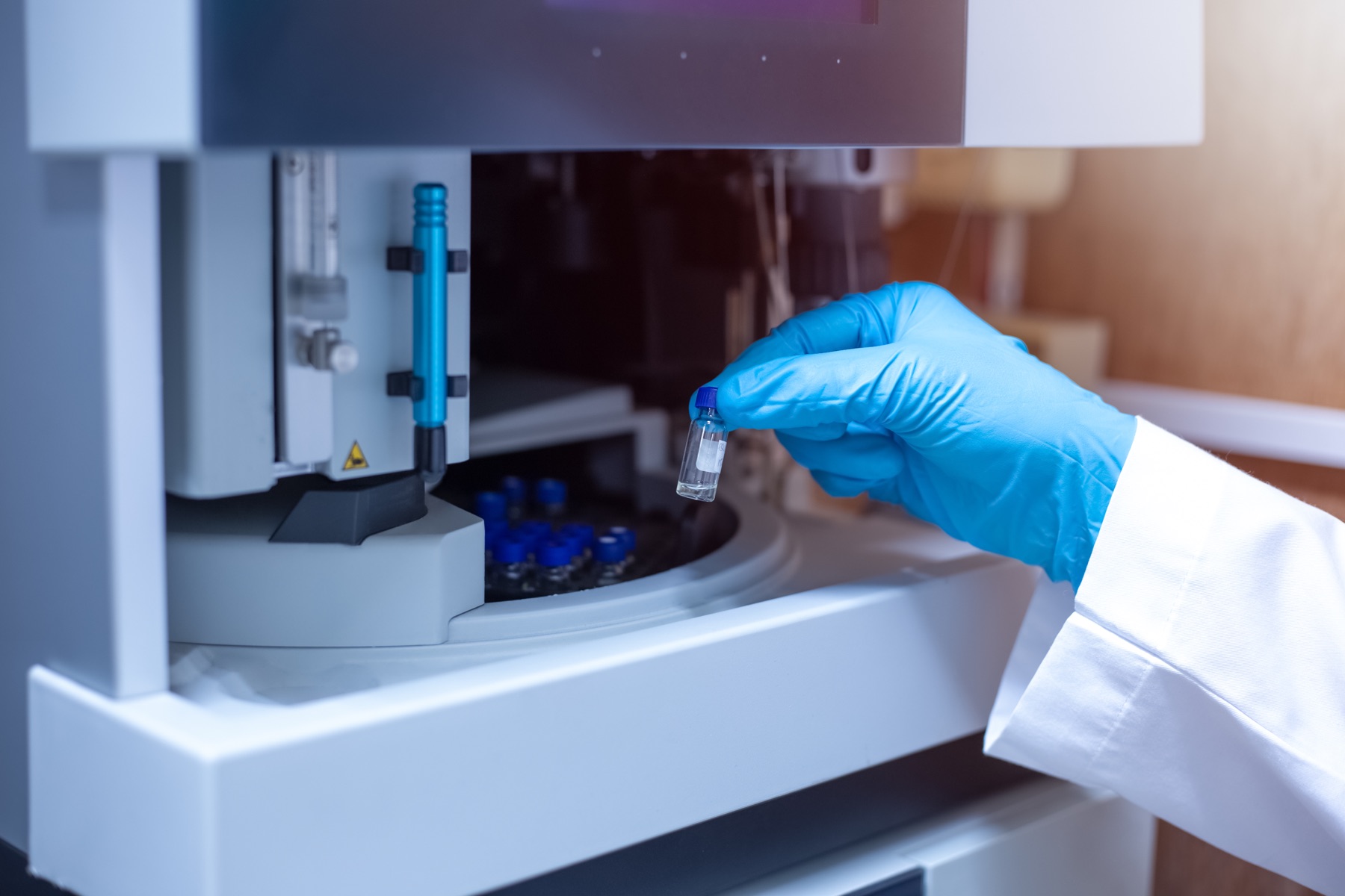 A blue-gloved hand lifting a sample vial from an LC/MS autosampler.